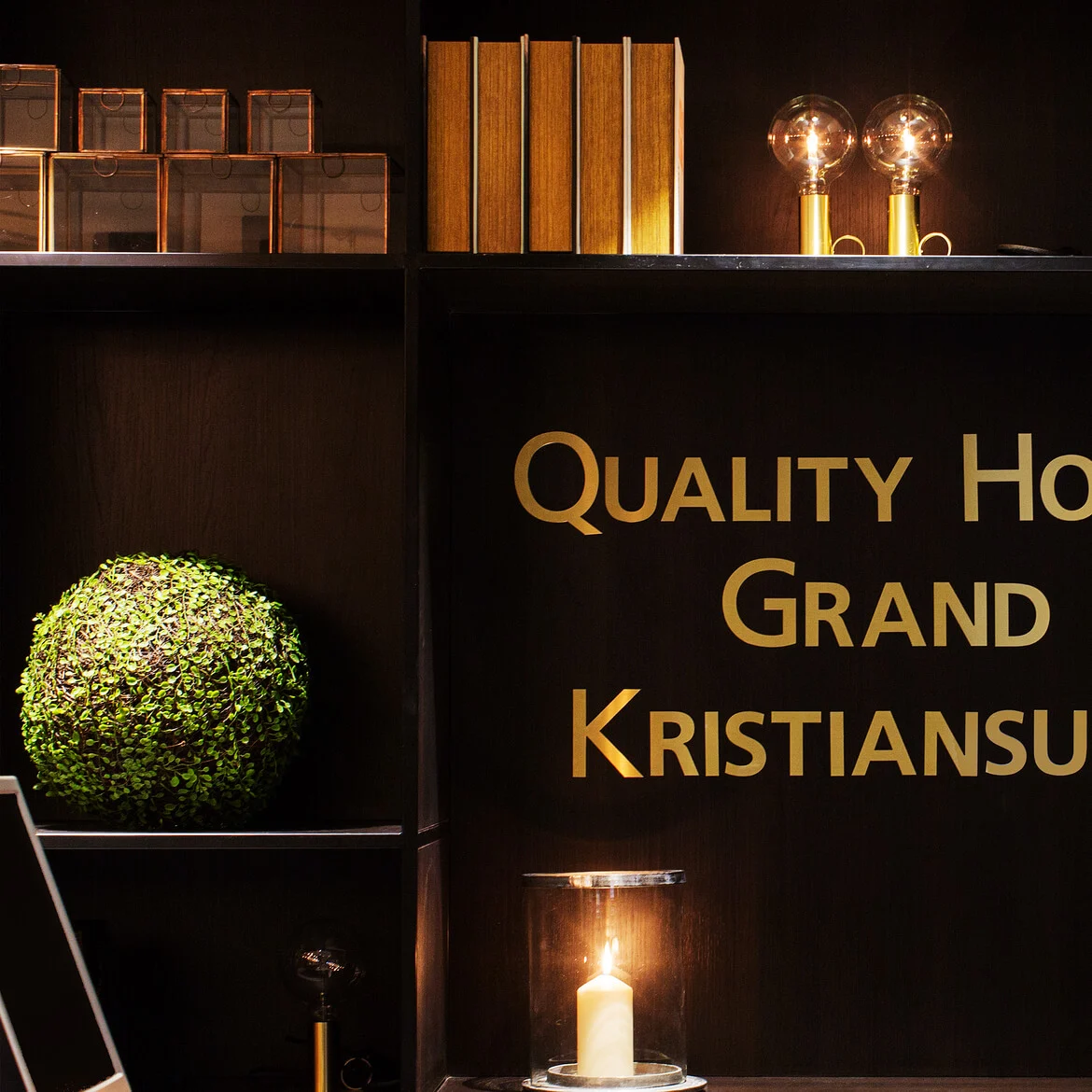 Quality Hotel Grand Kristiansund, tekst i gull bokstaver, dekorerer en mørk hylle. Bøker, runde grønne planter, to glassdekte pærer og et lys er plassert på omkringliggende hyller.