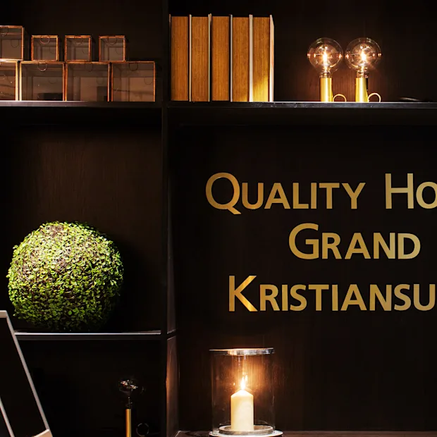 Quality Hotel Grand Kristiansund, text in gold letters, decorates a dark shelf. Books, round green plants, two glass-covered bulbs, and a candle are arranged on surrounding shelves.