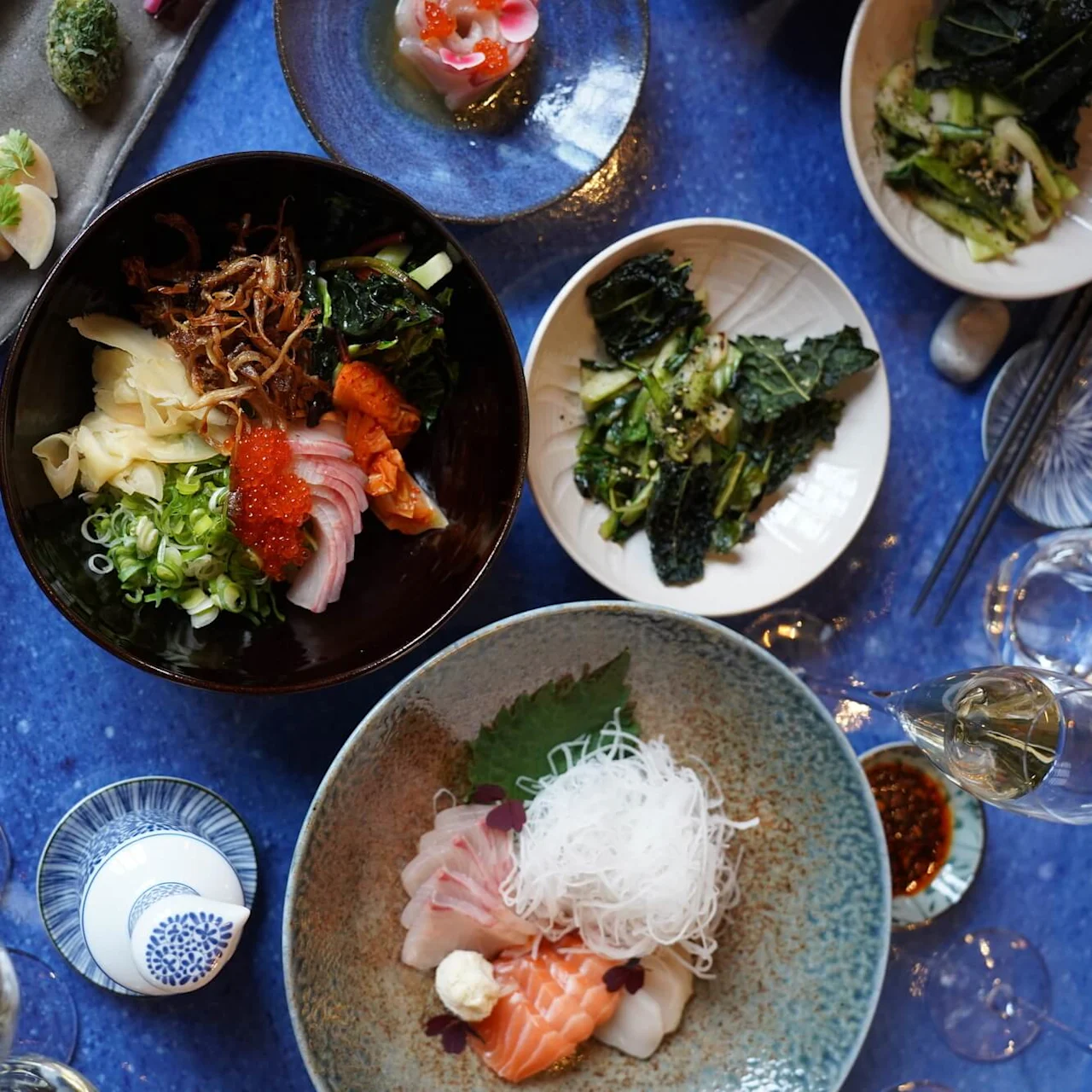 Erilaisia japanilaisia ruokia, kuten värikästä sushia ja tuoreita vihanneksia, aseteltuna sinisen pöytäliinan päälle, mukana misokeittoja ja kuplavesilaseja, luoden elegantin ruokailuympäristön.