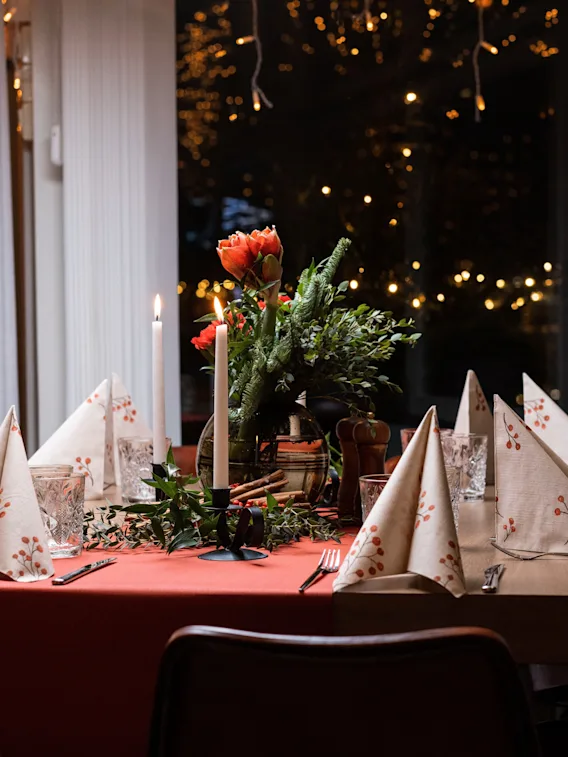 Elegant middagsbord, uppdukat med vikta servetter och ljus, omgivet av varma ljus; har en central blommig arrangemang med röda och vita blommor, i en mysig restaurangmiljö.