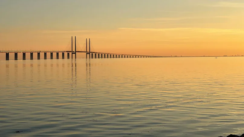 Bridge extends across calm water, reflecting a sunset sky with orange and blue hues, creating a serene atmosphere.