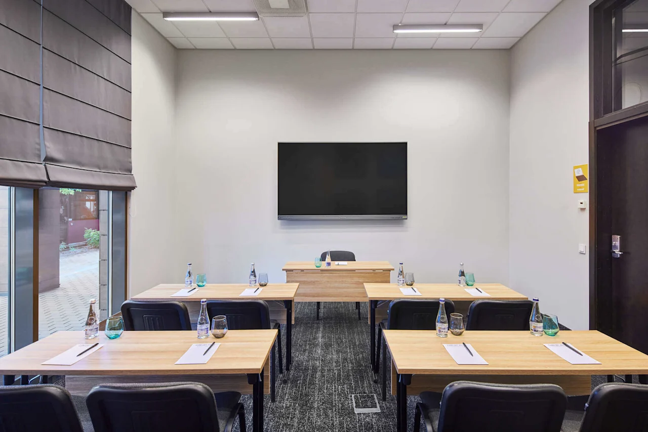 A conference room with a large screen on the wall, six desks arranged in rows, each with notepads, pens, and water bottles. Natural light comes through a window.