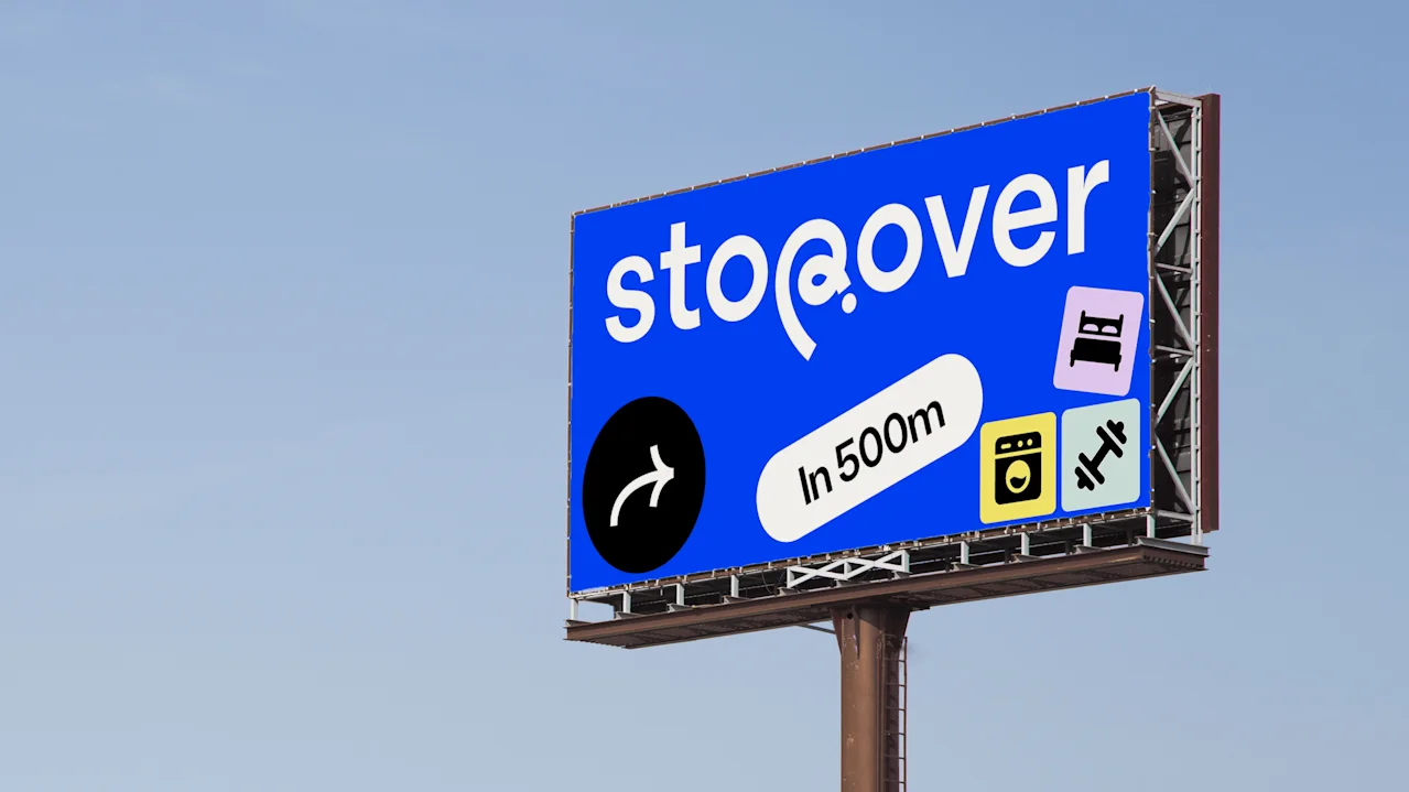 Mainostaulu näyttää "stopover" sinisellä taustalla. Kuvakkeet sängystä, pesukoneesta ja käsipainosta. Nuoli oikealle tekstillä "In 500m." Kirkasta taivasta vasten.