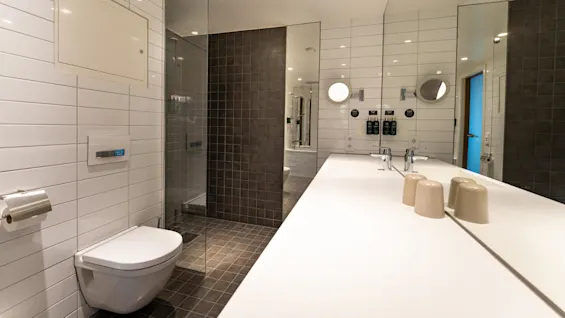 Et moderne badeværelse med væghængt toilet, glasbruser, lang bordplade med dobbeltvask, spejle og hylder med toiletartikler. Lys belysning og hvide flisevægge fuldender det elegante design.
