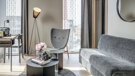 Grå soffa, runt soffbord med blommor och fåtölj är arrangerade i ett modernt vardagsrum. En golvlampa står bredvid stolen, nära stora fönster med stadsutsikt.
