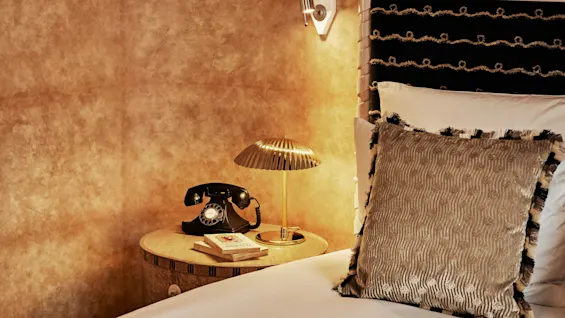 Et koselig soveromshjørne med en vintage roterende telefon på et rundt nattbord ved siden av en teksturert pute og en messinglampe, mot en varmtonet vegg.
