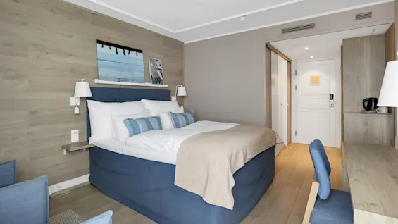 En pent seng med blå aksenter står i et moderne hotellrom. Trevegger, et lite skrivebord, stol og vannkoker kompletterer det minimalistiske interiøret. En hvit dør er litt åpen.