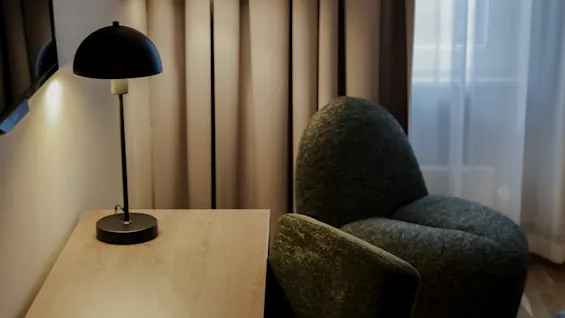 A desk with a black lamp sits next to a green chair in a softly lit room. A floor lamp illuminates the corner with draped curtains.