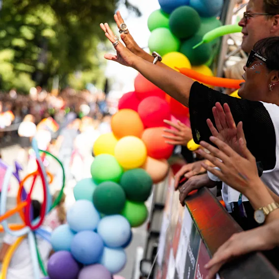 Folk jublar och vinkar från en paradvagn, med färgglada ballonger längs kanten. Den omgivande miljön är fylld av liv och rörelse, vilket antyder en festlig händelse.