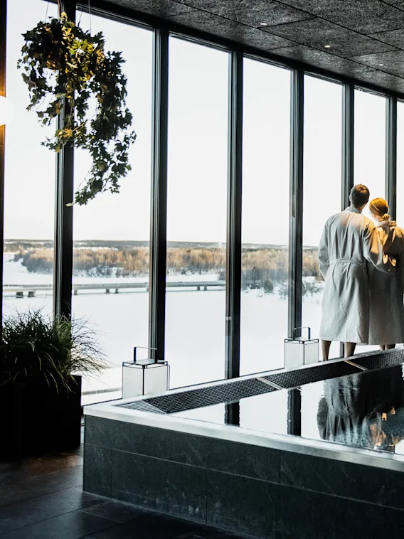 Ett par i vita badrockar står vid stora fönster och tittar ut på ett snötäckt landskap. De befinner sig i ett modernt spa med en reflekterande pool och loungestolar.