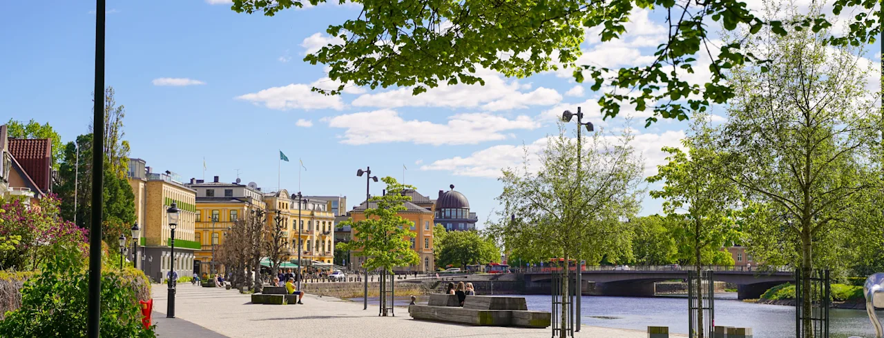 A sunny city promenade by a river with green trees and buildings in the background, perfect for att göra i Karlstad med barn.