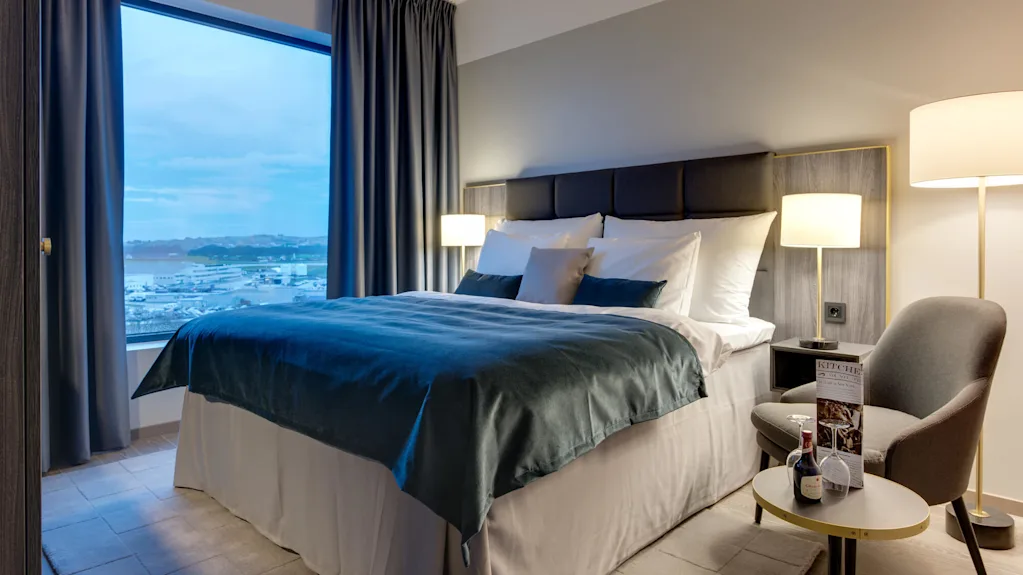 En stor säng med turkosa sängkläder står i ett modernt, välbelyst hotellrum. Ett fönster visar en naturskön vy över en marina och berg. Ett sidobord har en meny och en liten vinflaska.