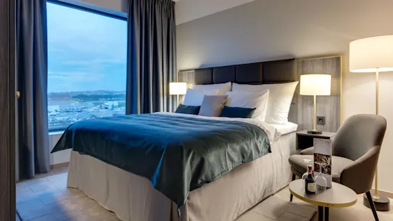 En stor seng med teal sengetøy står i et moderne, godt opplyst hotellrom. Et vindu avslører en naturskjønn utsikt over en marina og fjell. Et sidebord har en meny og en liten vinflaske.