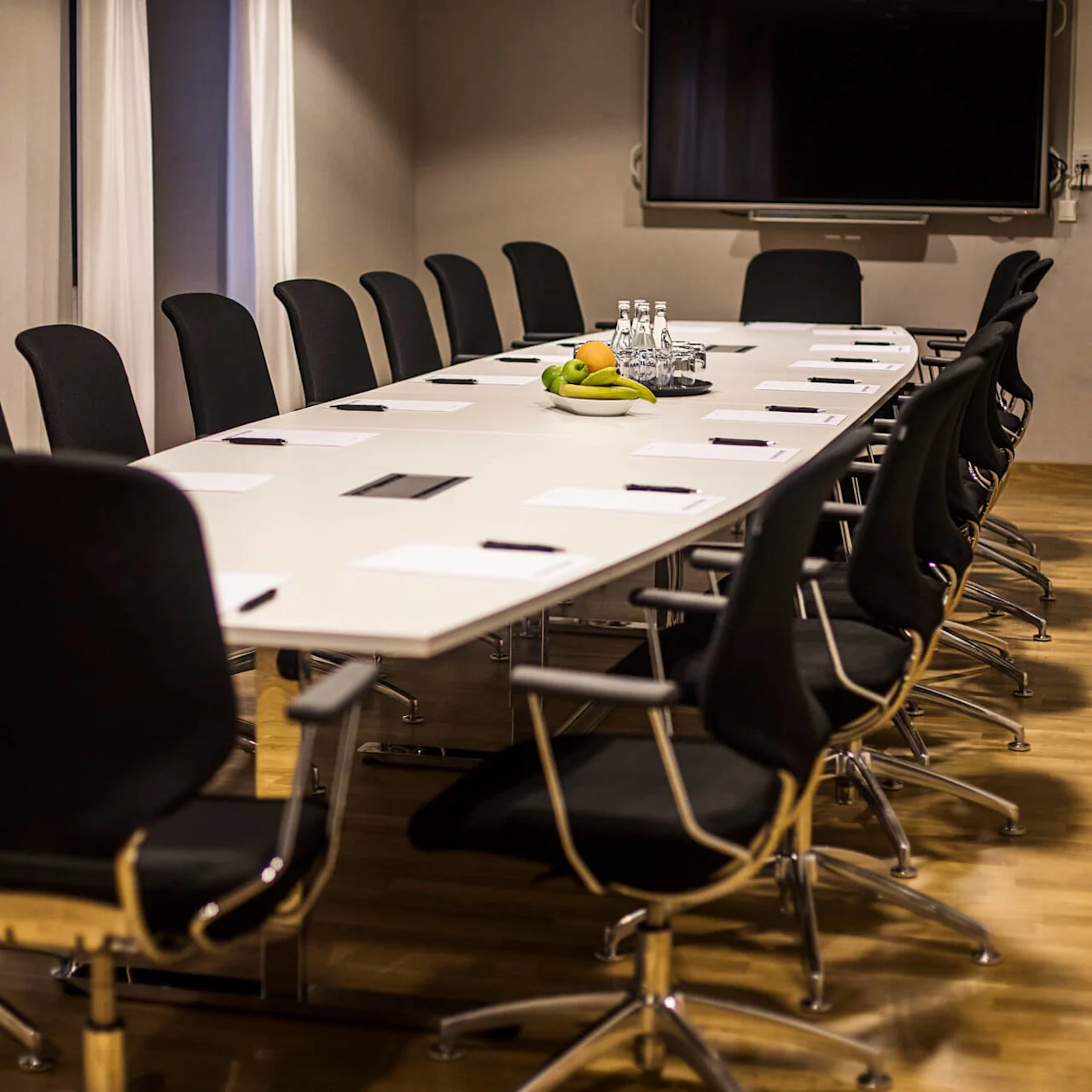 Empty boardroom with chairs around a long table, a fruit bowl in the center, a display screen on the wall.