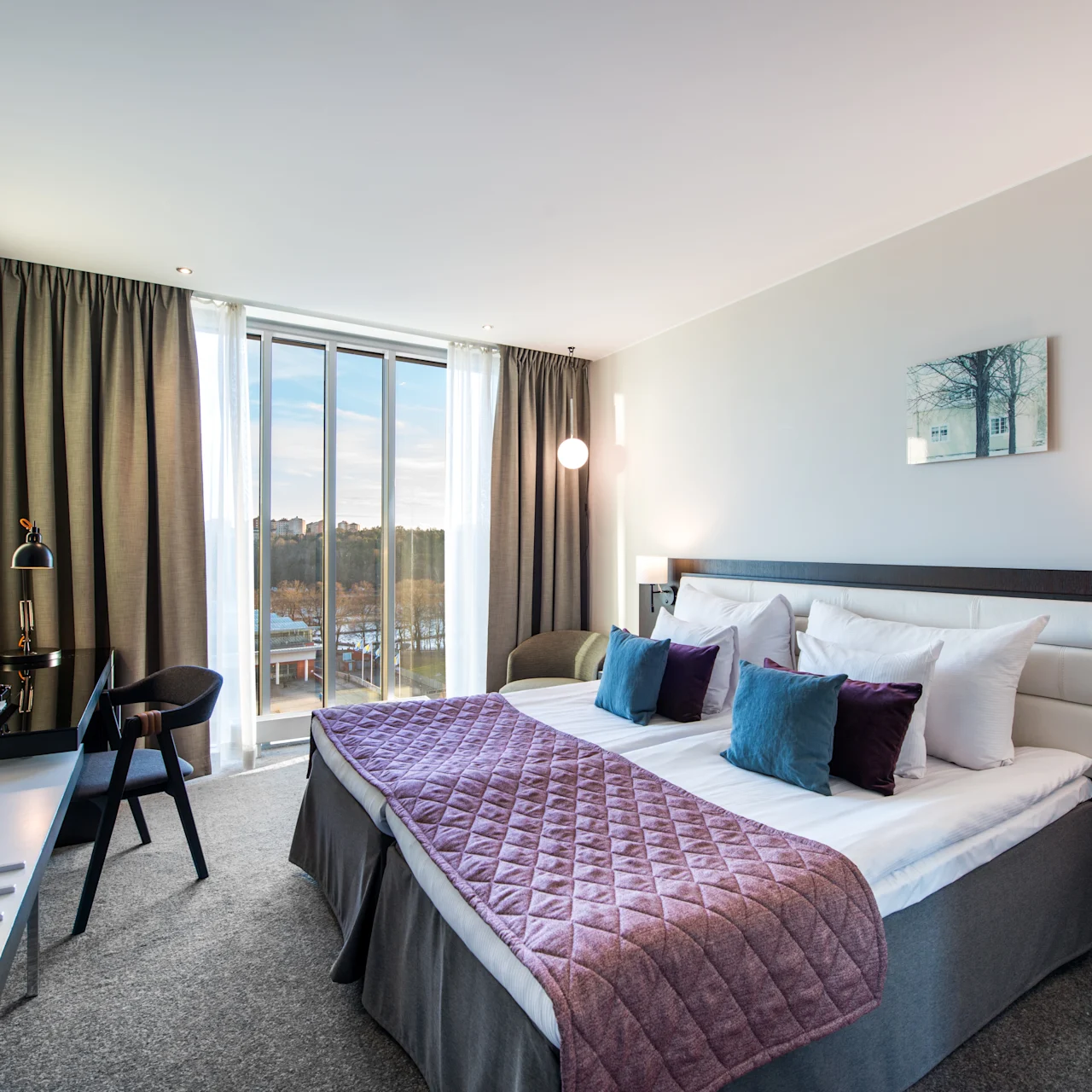 En säng med kuddar och ett kviltat överkast är centralt belägen i ett väl upplyst hotellrum med gardiner, ett skrivbord, stol, väggmonterad TV och ett stort fönster.