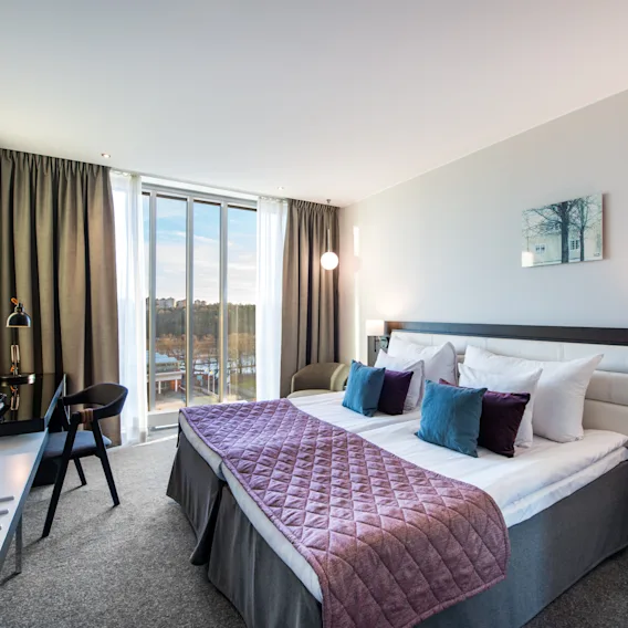 A bed with pillows and a quilted throw is centrally located in a well-lit hotel room with curtains, a desk, chair, wall-mounted TV, and a large window.