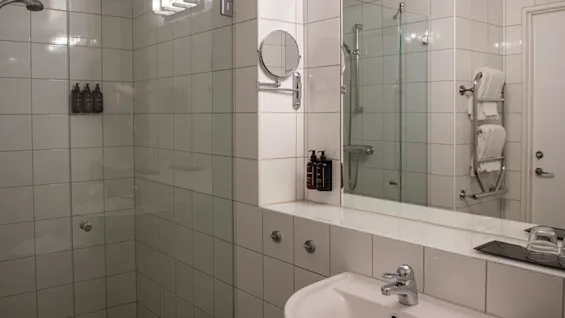 Sink in a tiled bathroom, equipped with a mirror, soap dispensers, and overhead lighting. Adjacent is a glass-walled shower and towel rack, in a minimalist style.