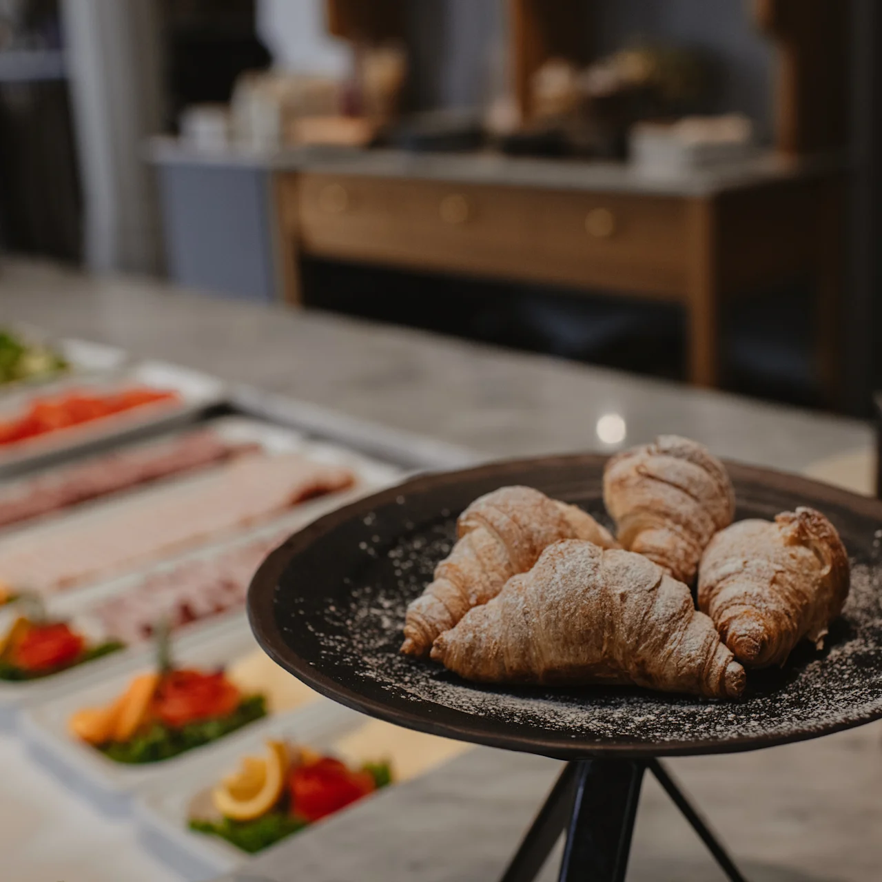 Croissanter ligger på ett svart fat beströdda med florsocker; i närheten visas skivade kött, ostar och grönsaker på ett buffébord i en restaurangmiljö.