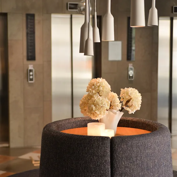 A gray circular sofa with white flowers and candles rests in a lobby. Behind it, three elevator doors are visible, framed by neutral-toned walls and illuminated by overhead pendant lights.