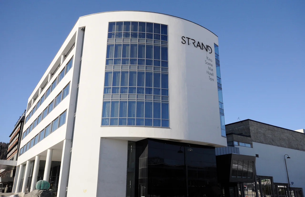 A modern multi-story building with wide glass windows curves upwards under a clear blue sky. "STRAND" is prominently displayed on the facade with smaller text: "Sola Hotel Spa."