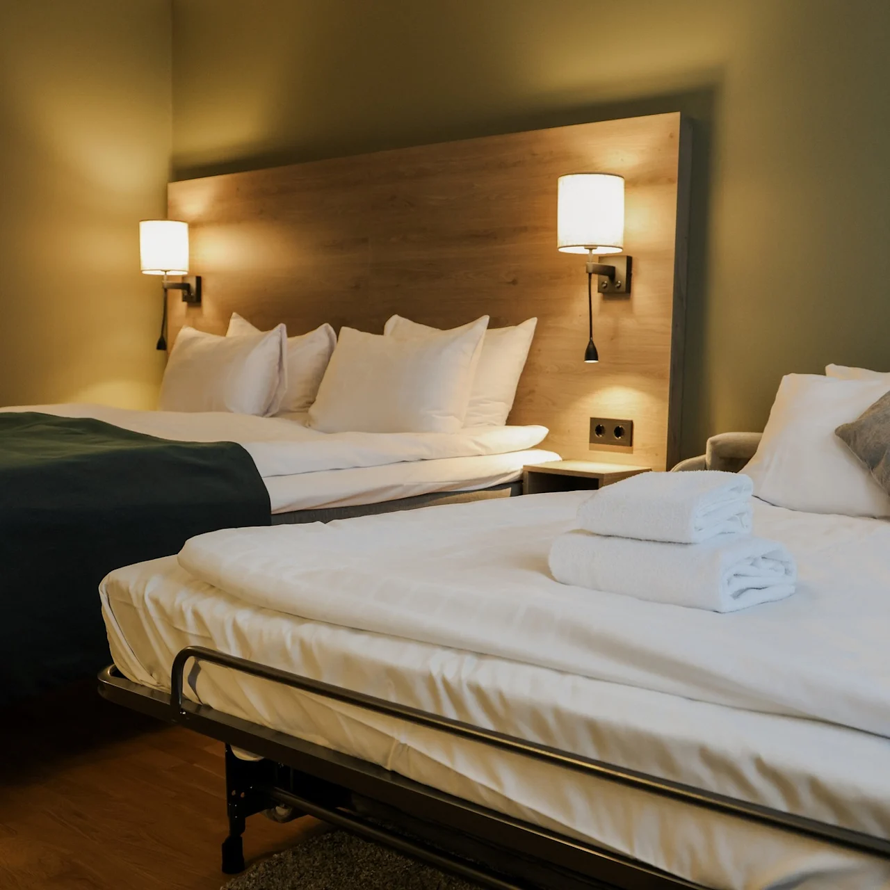 A neatly arranged hotel room features two beds, one with a green blanket and plush pillows, and a smaller bed with folded towels; wooden bedside tables and framed artwork enhance the cozy ambiance.