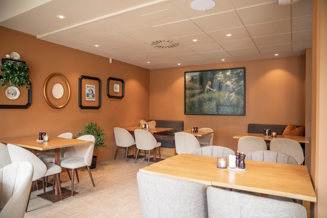 Tables and chairs arranged in a cozy café setting, surrounded by warm, earthy-colored walls adorned with framed art. Ambient lighting creates a relaxed atmosphere.