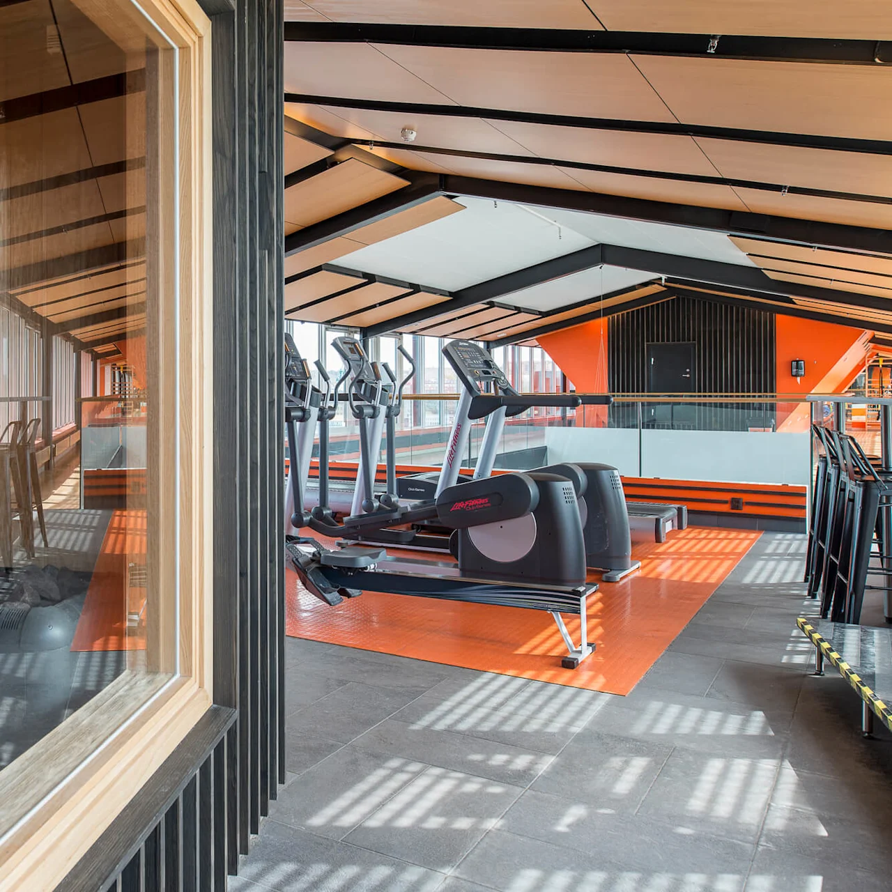 Treadmills align in a modern gym. Action: sunlit through large windows. Context: adjacent sauna room with wooden benches and a heater. No visible text.
