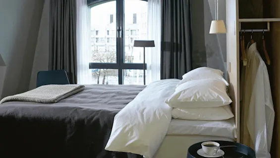 A bed in a modern room with gray bedding, placed near a large arched window. A cup and saucer sit on a side table, with a chair and wardrobe nearby.
