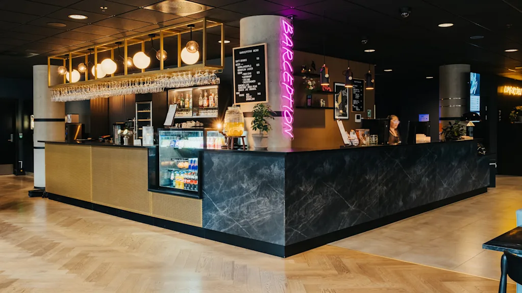 Bar med hængende glas og oplyste skilte, placeret i en stilfuld, moderne indretning. Neon "BARCEPTION" skilt er fremtrædende. Menu: SOFT DRINKS 39, SMOOTHIES/FRAPPÉS 59, SPECIAL-LOLAS LICOR (ALCOHOL).