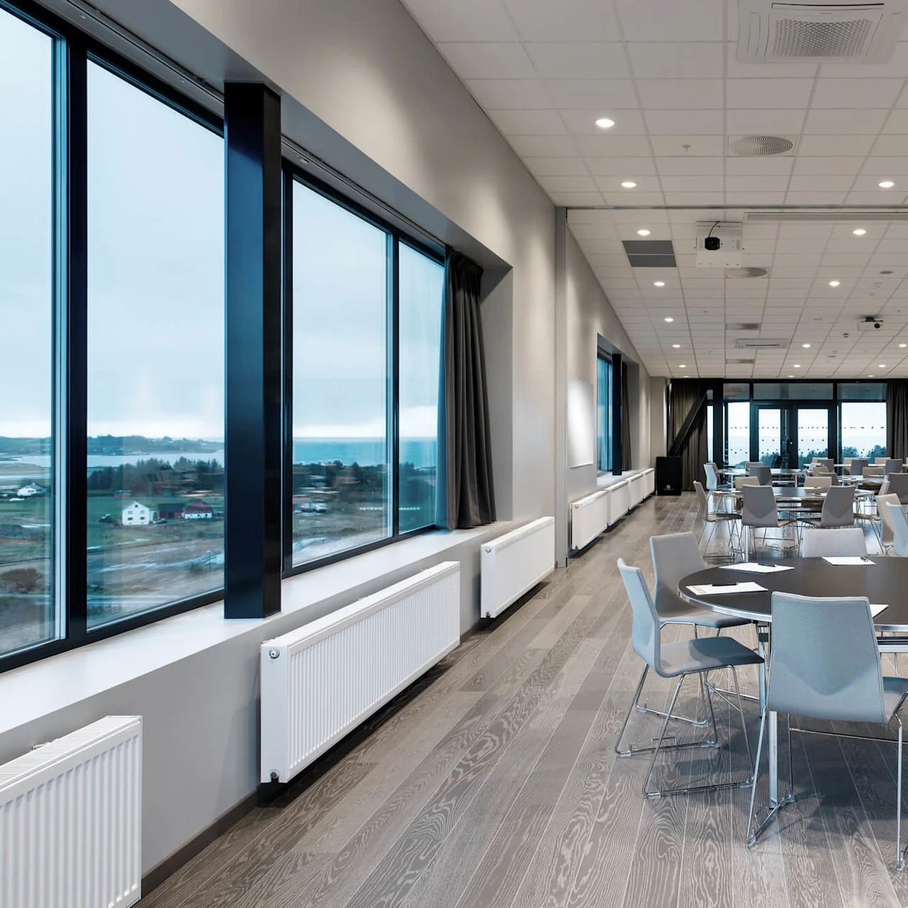 Et konferanserom med runde bord og stoler er ordnet pent, klart for et møte. Store vinduer på én side gir utsikt over et overskyet landskap.