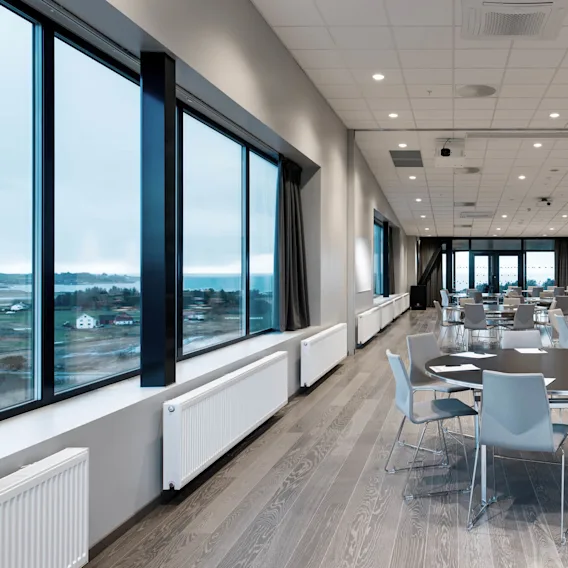Et konferanserom med runde bord og stoler er ordnet pent, klart for et møte. Store vinduer på én side gir utsikt over et overskyet landskap.