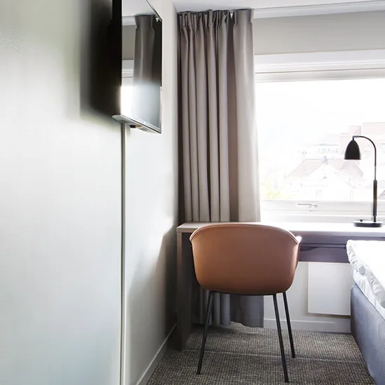 En seng står pænt i et moderne hotelværelse med et vægmonteret TV overfor. Et skrivebord og en stol er placeret ved siden af et stort vindue med lukkede gardiner.