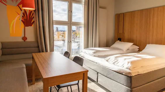 A bed with white linen is beside a wooden table and chairs in a room with a large window and colorful wall design.