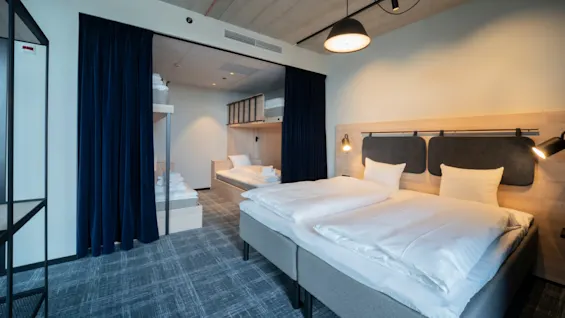 Moderni hotellihuoneessa on kaksi siististi pedattua sänkyä valkoisilla lakanoilla. Vieressä on toinen huone, jossa on kerrossängyt, osittain piilossa tummansinisten verhojen takana. Katto on näkyvillä kanavilla.