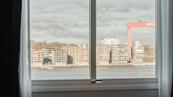 A window frames a riverside view with apartment buildings, a crane labeled "ERIKS," and a cloudy sky.