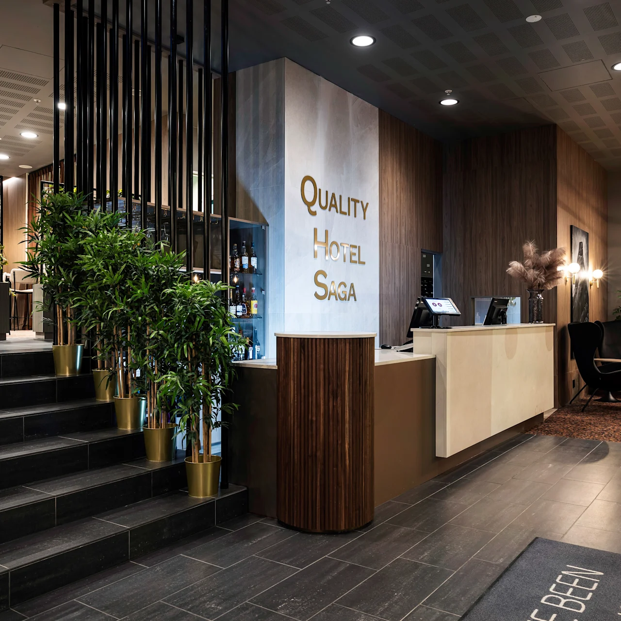 Reception desk at "Quality Hotel Saga" with modern design, plants, and decor elements like a round mirror and stylish lighting. Stepped entrance leads to an inviting seating area.