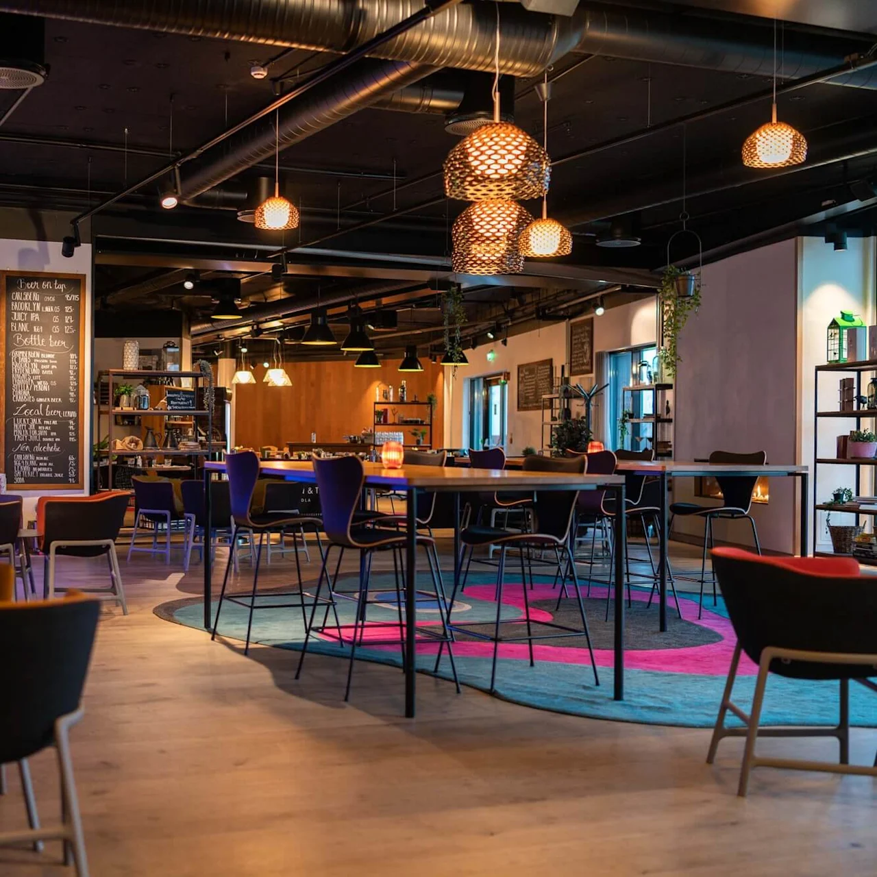 En rymlig kafé har moderna möbler och konstnärlig inredning. Stolar omger träbord under varm belysning. En stor svart tavla på väggen listar menyer vilket förstärker den mysiga atmosfären.