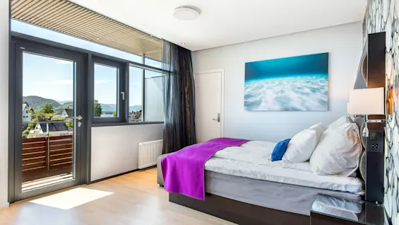 Et moderne soverom med en seng med lilla og hvitt sengetøy, ved siden av et vindu med utsikt over hus og åser. Et havlandskap dekor pryder veggen.