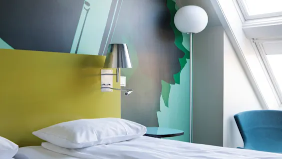 A neatly made bed with white sheets is next to a green wall with a mounted chrome lamp; a modern chair and a floor lamp stand near a bright window.