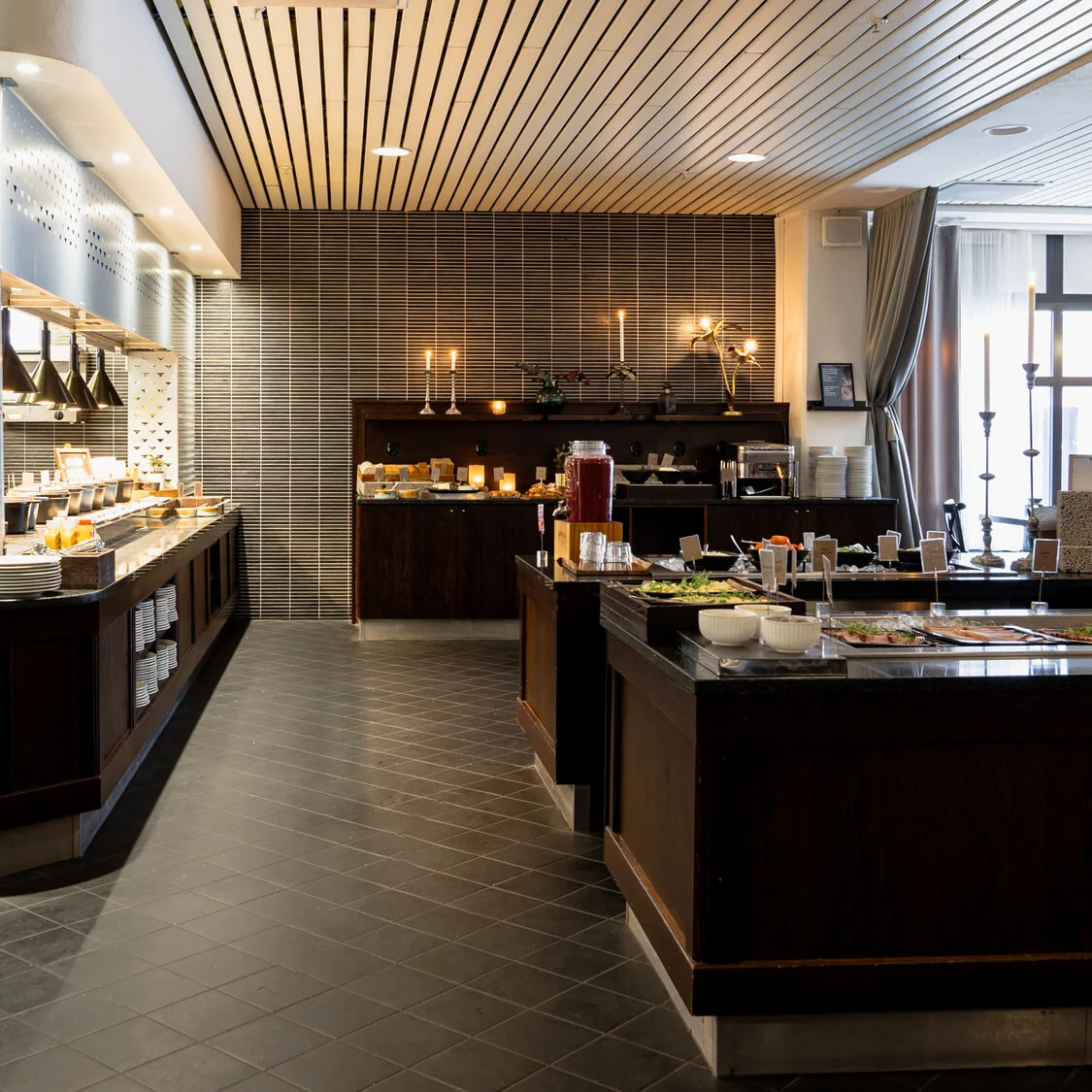 En buffetopstilling med pænt stablede tallerkener, der byder på forskellige morgenmadsprodukter, salater og drikkevarer. Rummet har et moderne design med store vinduer og stearinlys som stemningsskabere.