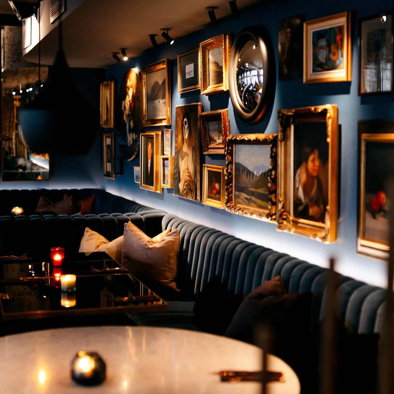 Wall displays various framed artworks illuminated by spotlights. Below, plush blue seating surrounds dark tables with lit candles, creating a cozy, intimate lounge atmosphere.