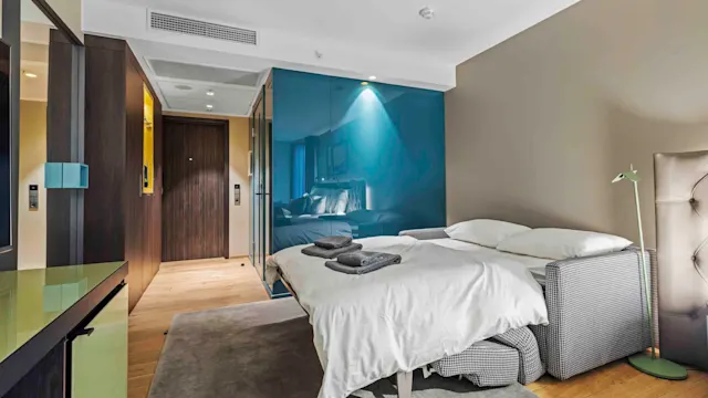 En seng med hvitt sengetøy er pent oppredd, vendt mot en døråpning. Brettede håndklær ligger oppå. Et moderne rom med tregulv, blå glassdetaljer og dempet belysning.