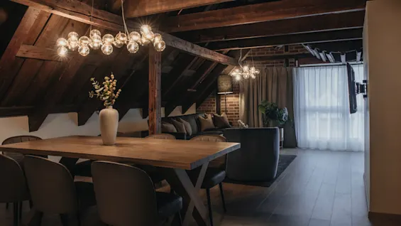 Et hyggeligt loftsopholdsrum med spisebord og loungeområde, med synlige træbjælker og varm belysning.