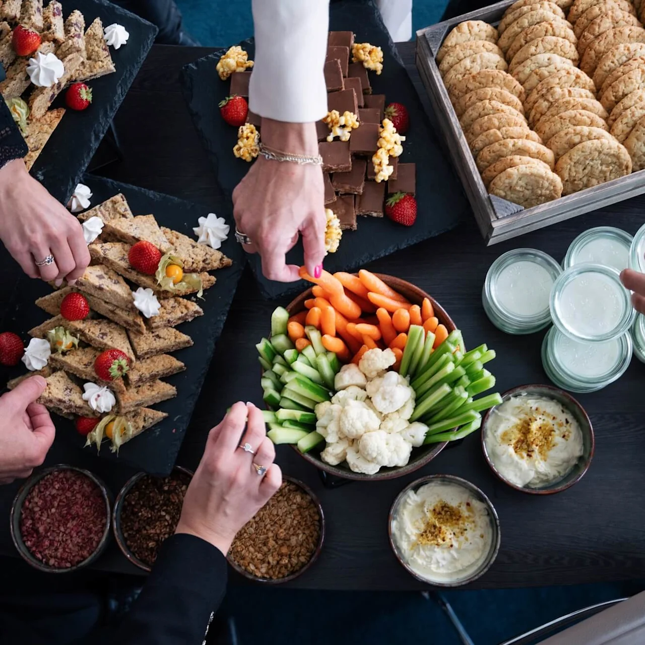 Platters of assorted fruits, cookies, and vegetables with dips are arranged on a table. Multiple hands are reaching for food. Plates and glasses are placed around the spread.