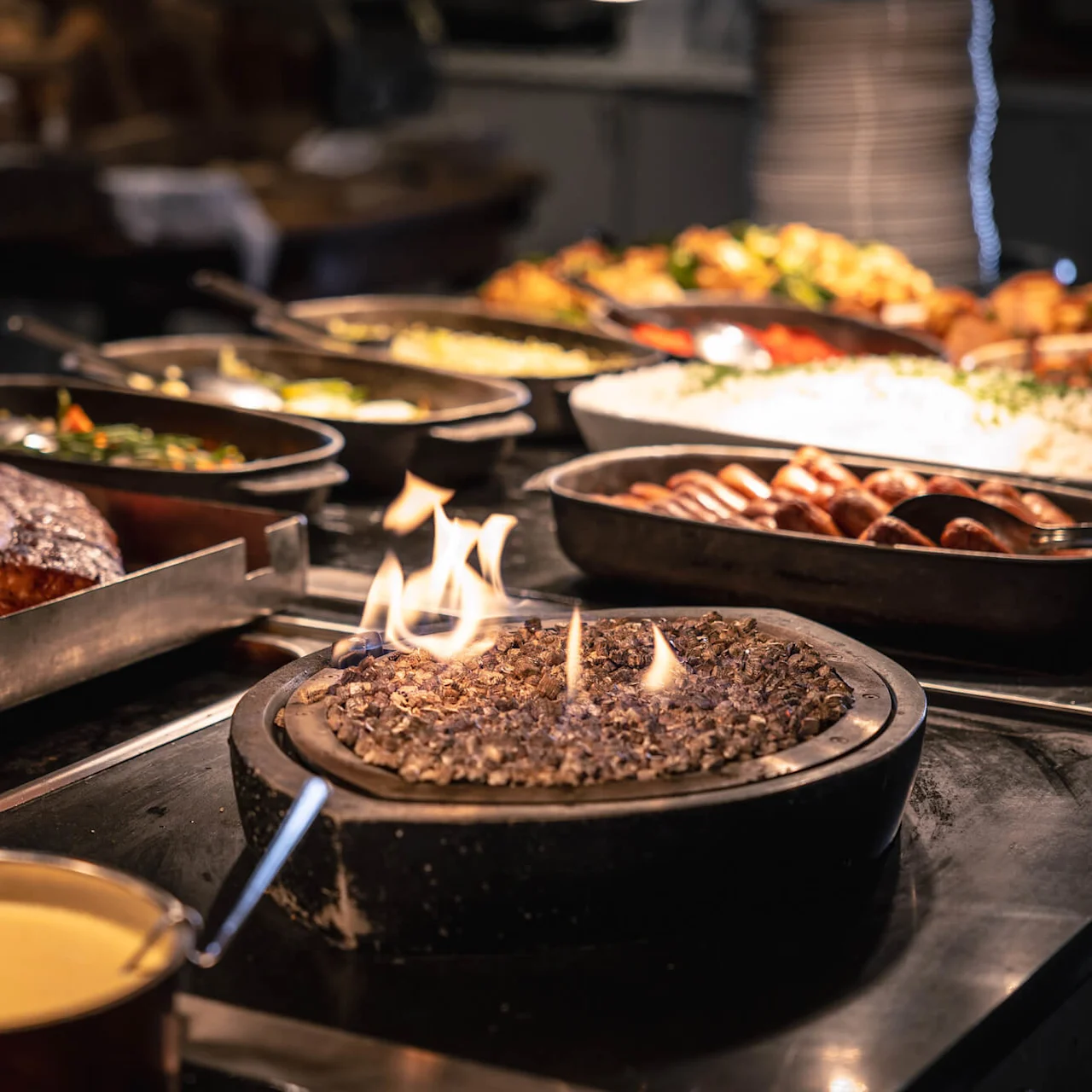 Buffetdisplay med stekt kjøtt og forskjellige tilbehør i serveringsbrett. Flammer holder en rett varm. Tallerkener og bestikk klare for servering i en varm, innbydende atmosfære.