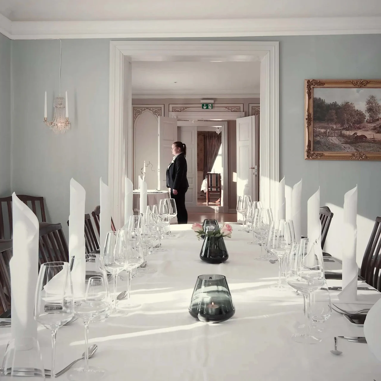 Et langt, elegant dækket spisebord med hvid dug, glas og foldede servietter; en person står i baggrunden ved store døre i et smukt dekoreret rum.