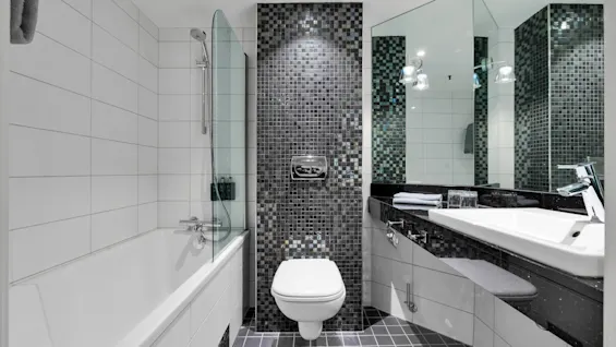 Toilet på en mosaikvæg, flankeret af et badekar med glasskærm til venstre og en vask med spejl og håndklæde til højre. Moderne badeværelsesdesign.