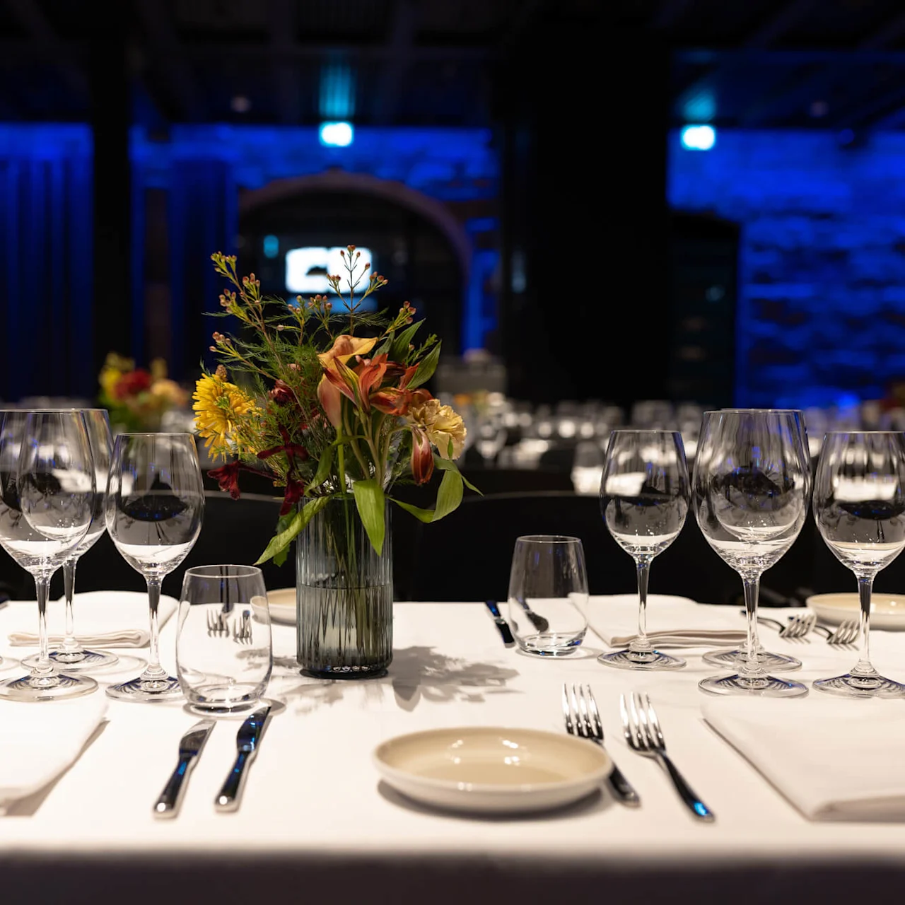 A table elegantly set with wine glasses and flower arrangements, in a dimly-lit, blue-accented banquet hall.