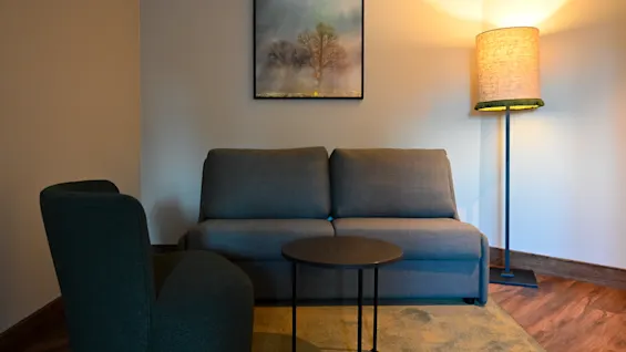 Et hyggeligt hotelværelseshjørne med en grå sofa, lænestol, sofabord og varm belysning, ideelt til afslapning.