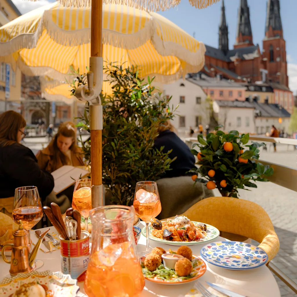 Outdoor dining scene with people enjoying food and drinks under a yellow umbrella, a cathedral in the background.